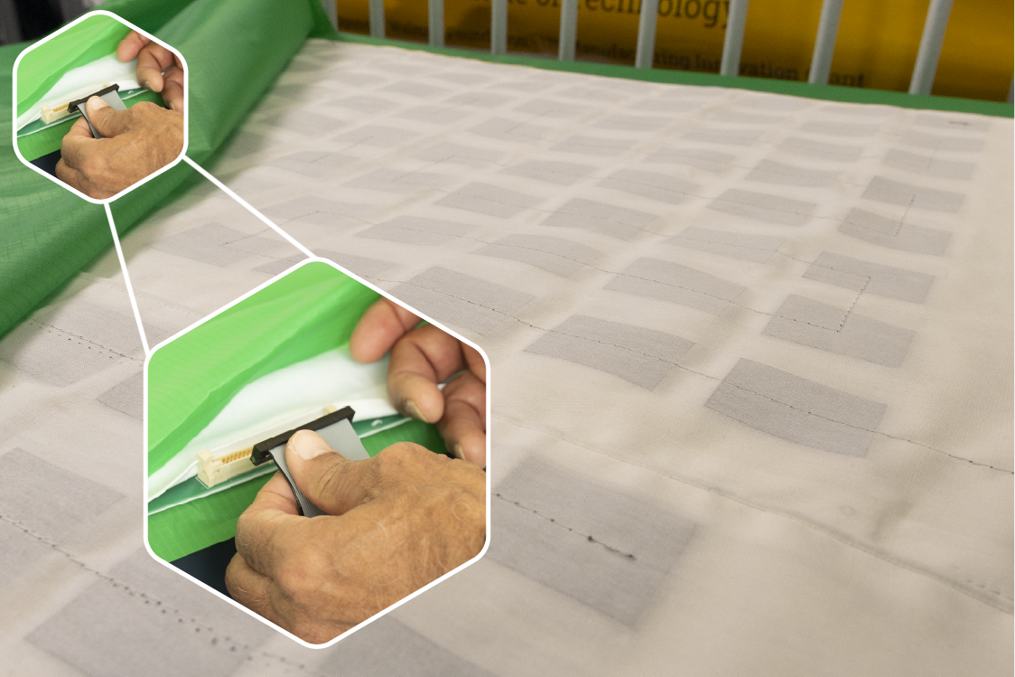 A close-up view of a crib mattress covered with white pressure‑sensing fabric featuring a grid pattern, with an inset image showing a pair of hands connecting a singular cable.