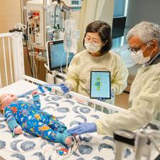 Two adults wearing protective gowns and gloves stand beside a hospital crib, using a tablet device while examining an infant lying on the mattress as medical equipment and monitors surround the crib.